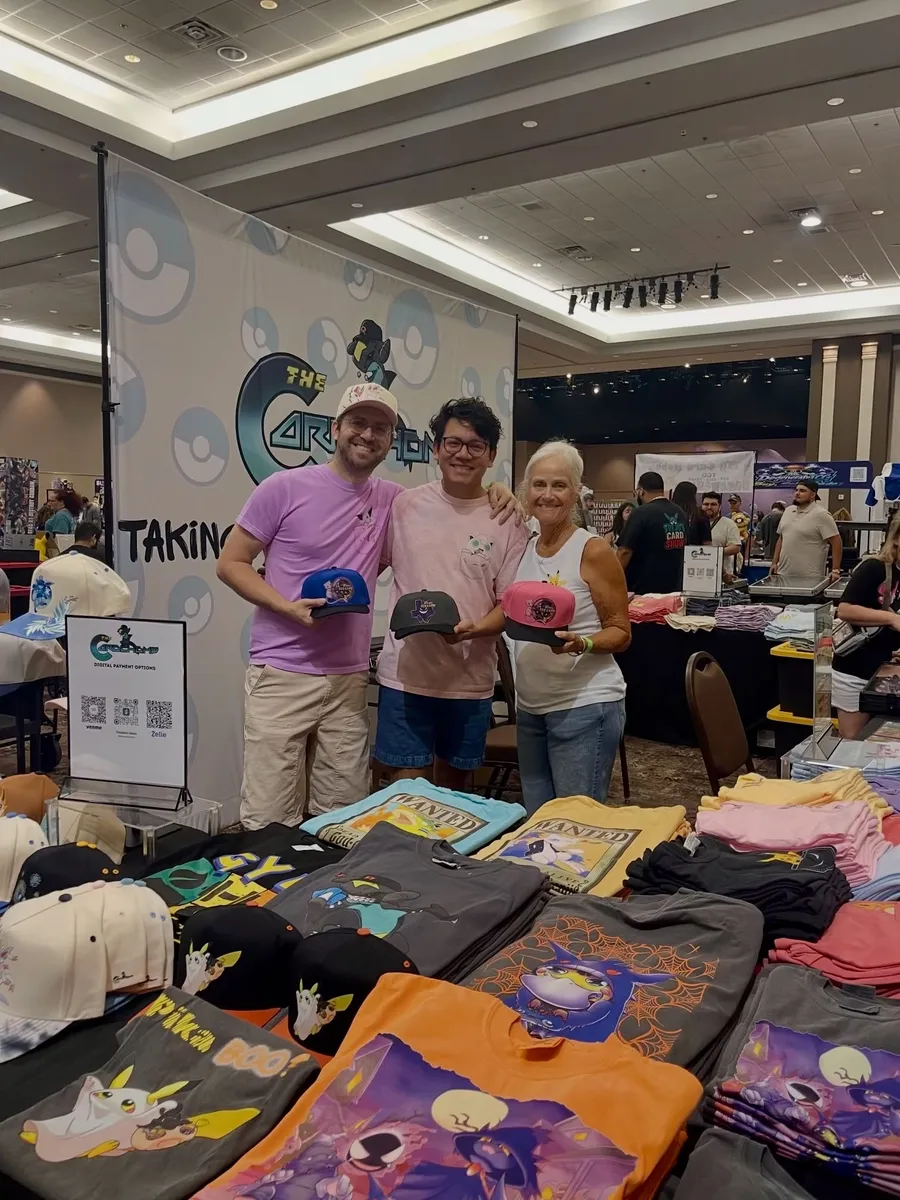 Three people holding Texas TCG Expo hats behind a table of Pokémon shirts